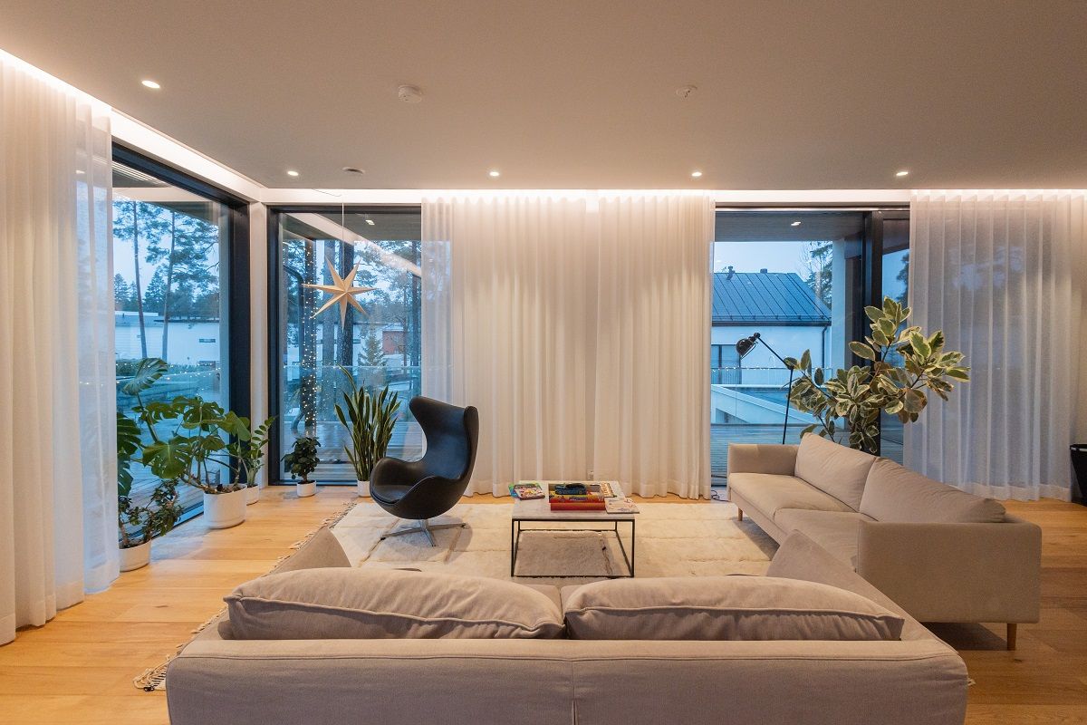 Il soggiorno è illuminato da faretti a incasso e indirettamente da strisce LED nella scanalatura del soffitto che illuminano le tende da giorno, bloccando la vista dall'esterno all'interno durante le ore di buio.